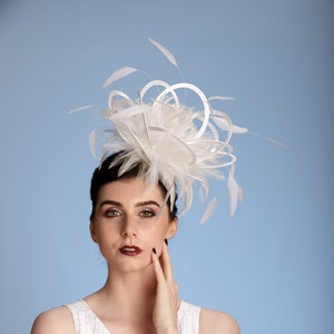 May include: A white fascinator with feathers and looped details. The headpiece is worn by a person with dark hair and a white dress. The background is a solid light blue.