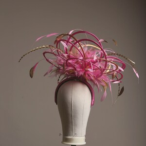 以下が含まれることがあります： ピンクとゴールドのヘッドピース、ヘアバンドベース。ピンクの羽根、ゴールドのアクセント、装飾が特徴です。マネキンの頭に飾られています。