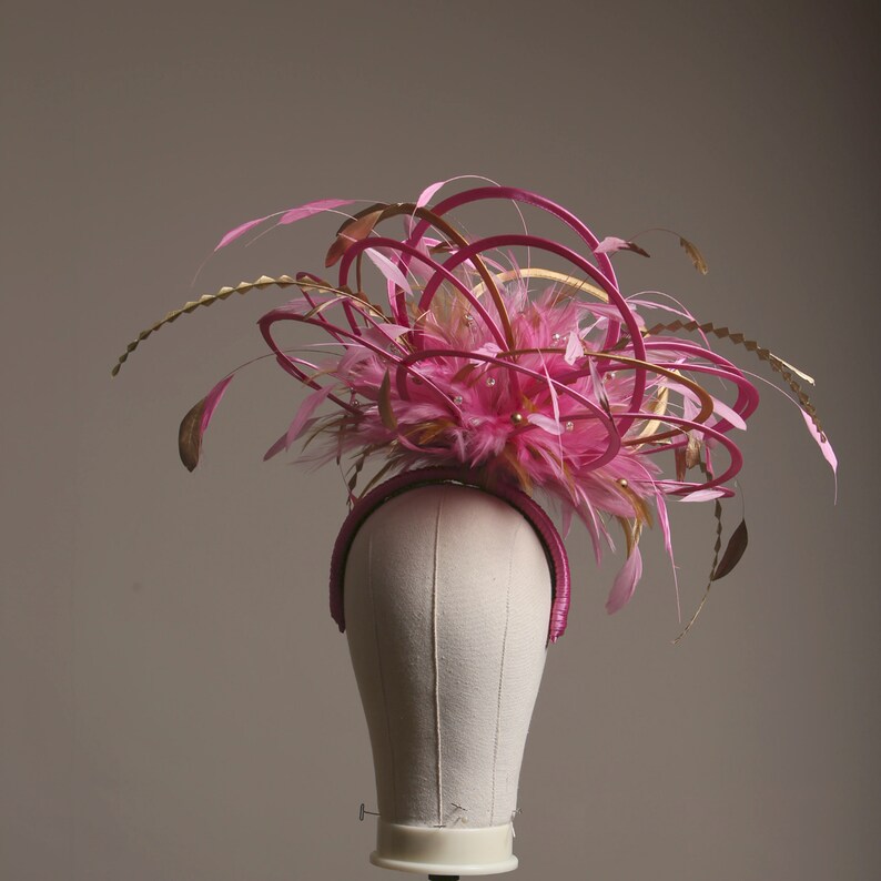 以下が含まれることがあります： ピンクとゴールドのヘッドピース、ヘアバンドベース。ピンクの羽根、ゴールドのアクセント、装飾が特徴です。マネキンの頭に飾られています。