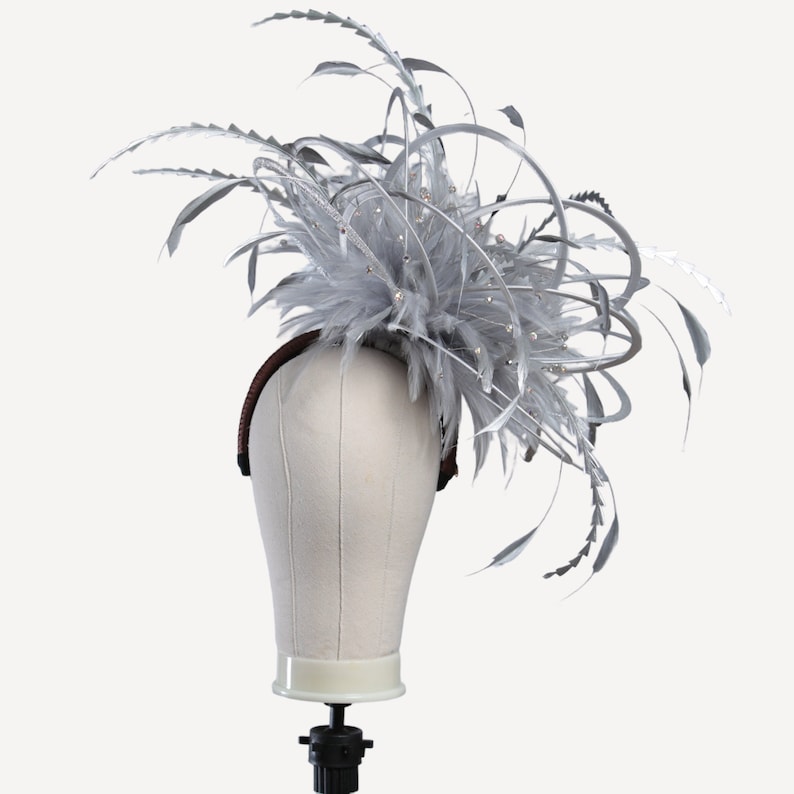 May include: A silver-colored fascinator with feathers and decorative accents. The headpiece features a headband base with a cluster of feathers and decorative elements, perfect for special occasions.