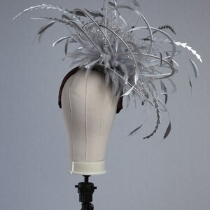 May include: A silver headpiece with feathers and decorative accents. The headpiece is displayed on a mannequin head. The headpiece features silver feathers, metallic accents, and small, sparkling embellishments. The base of the headpiece is a brown headband.
