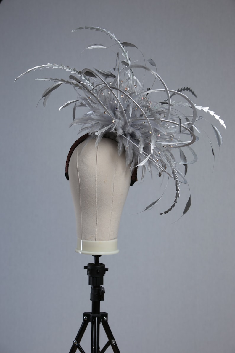 May include: A silver headpiece with feathers and decorative accents. The headpiece is displayed on a mannequin head. The headpiece features silver feathers, metallic accents, and small, sparkling embellishments. The base of the headpiece is a brown headband.