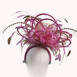 以下が含まれることがあります： ピンクとゴールドのヘッドピース。羽、ループ、装飾が施されています。ヘッドバンドは濃いピンク色で、デザインはピンクの羽と装飾的なアクセントのクラスターが特徴です。ヘッドピースは白いマネキンの頭に展示されています。