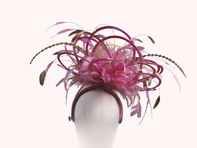 以下が含まれることがあります： ピンクとゴールドのヘッドピース。羽、ループ、装飾が施されています。ヘッドバンドは濃いピンク色で、デザインはピンクの羽と装飾的なアクセントのクラスターが特徴です。ヘッドピースは白いマネキンの頭に展示されています。