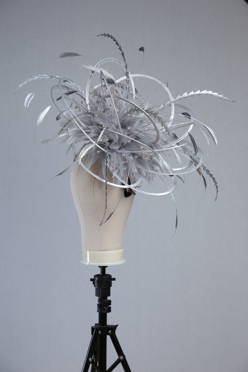 May include: A silver headpiece with feathers, metallic swirls, and sparkling embellishments. The headpiece is displayed on a mannequin head. This elegant accessory is suitable for special occasions.