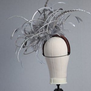 May include: A silver headpiece with feathers and decorative accents. The headpiece is mounted on a mannequin head. The headpiece has a brown headband and is adorned with silver feathers and sparkling embellishments.