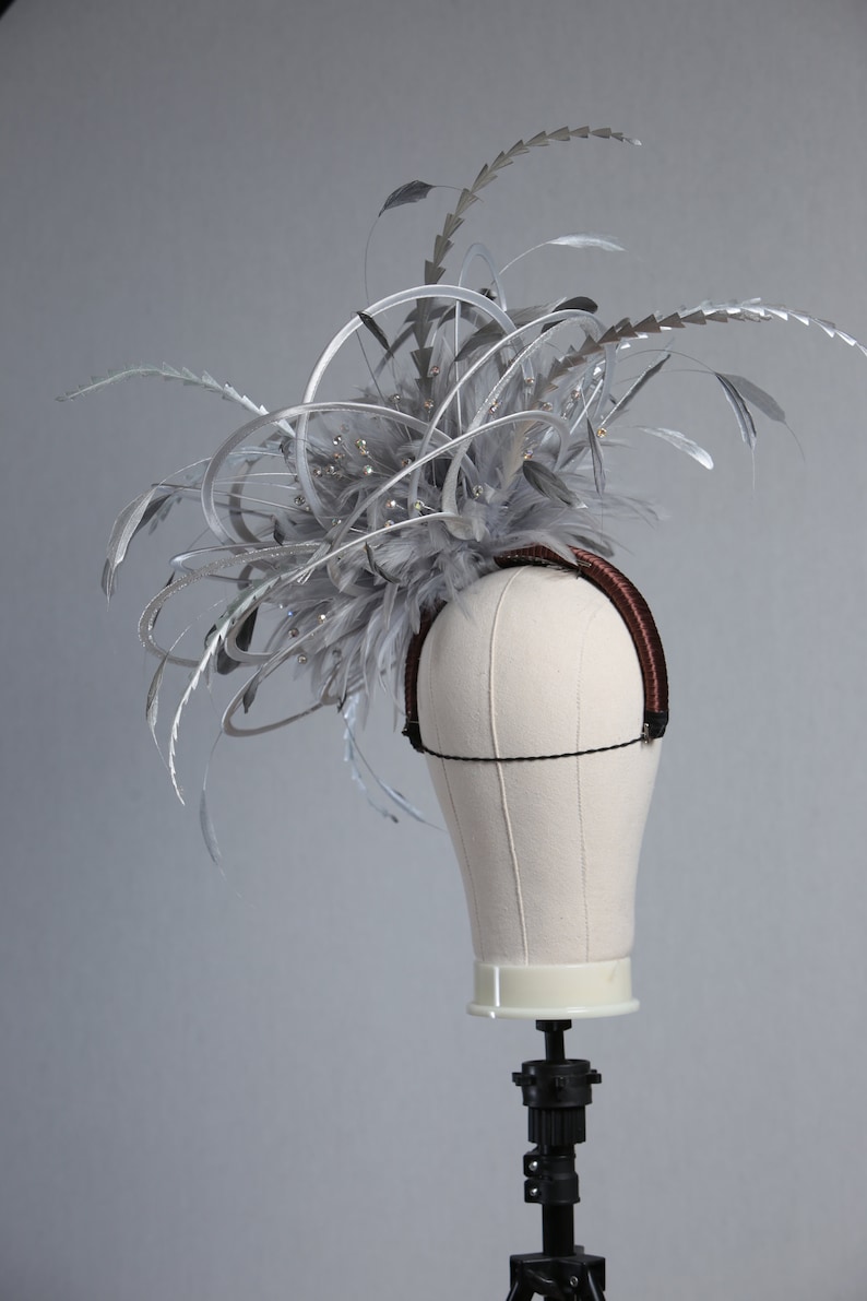 May include: A silver headpiece with feathers and decorative accents. The headpiece is mounted on a mannequin head. The headpiece has a brown headband and is adorned with silver feathers and sparkling embellishments.