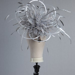 May include: A silver headpiece with a cluster of gray feathers, accented with small crystals and silver wire loops. The headpiece is displayed on a mannequin head, showcasing its design and structure. This accessory is suitable for special occasions.