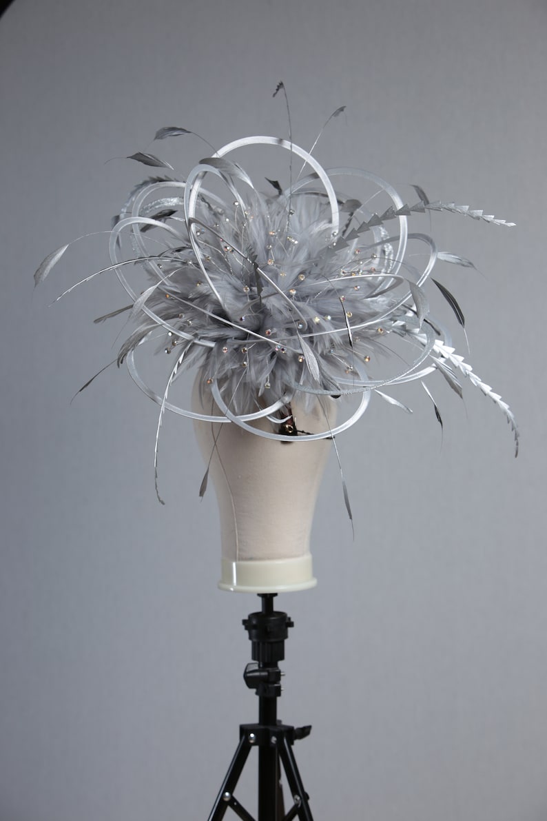 May include: A silver headpiece with a cluster of gray feathers, accented with small crystals and silver wire loops. The headpiece is displayed on a mannequin head, showcasing its design and structure. This accessory is suitable for special occasions.