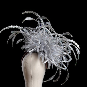 May include: A silver and gray fascinator with feathers and decorative metal accents. The headpiece features a cluster of gray feathers, silver metal swirls, and sparkling crystal embellishments. The base is a brown headband.