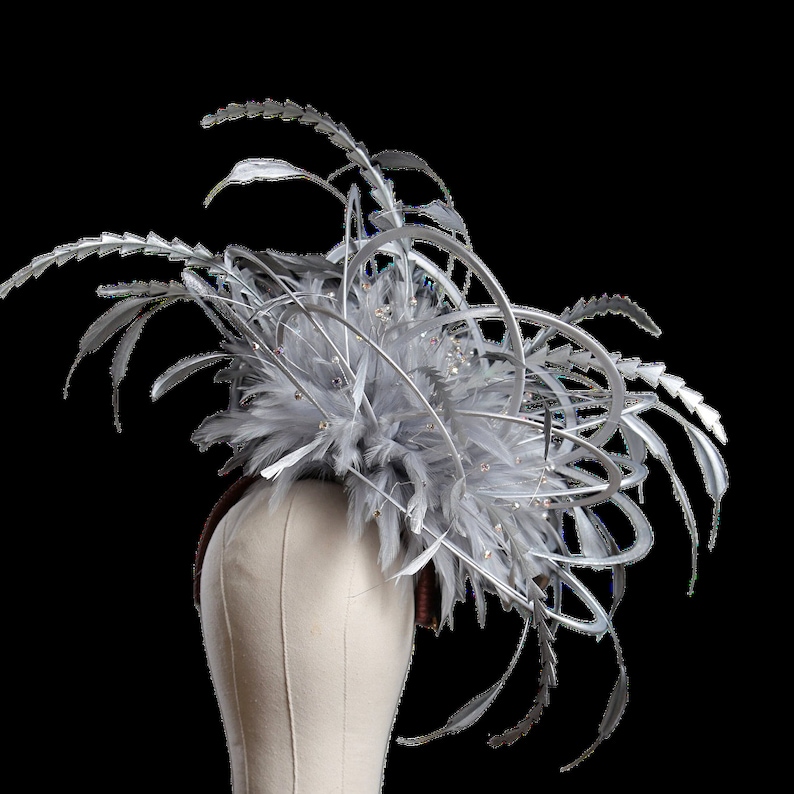 May include: A silver and gray fascinator with feathers and decorative metal accents. The headpiece features a cluster of gray feathers, silver metal swirls, and sparkling crystal embellishments. The base is a brown headband.