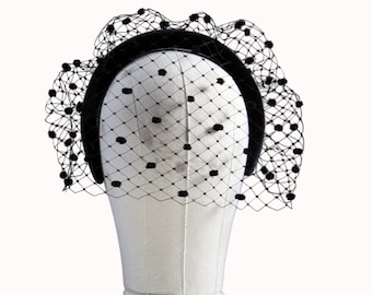 May include: A black headband with a velvet finish, adorned with a delicate black net veil. The veil features a grid pattern with evenly spaced black polka dots. The accessory is displayed on a mannequin head.