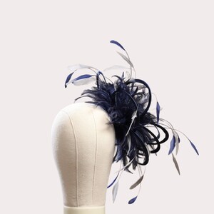 May include: A navy blue and white feather fascinator. The headpiece features a cluster of navy blue feathers, accented with white and navy blue feathers. The fascinator is mounted on a cream-colored mannequin head.