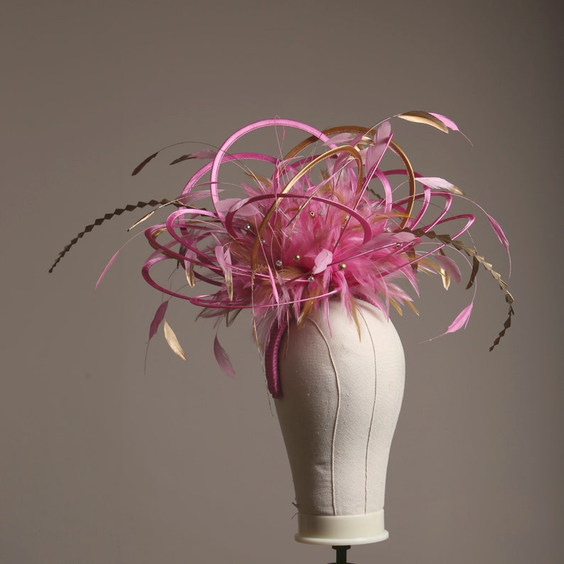 以下が含まれることがあります： 羽と装飾的なアクセントが付いたピンクとゴールドのファシネーター。ヘッドピースは、ピンクの羽の中央クラスター、金とピンクのループとアクセントで囲まれています。小さな透明な装飾がデザイン全体に散らばっています。