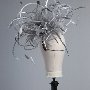 May include: A silver headpiece with feathers and sparkling embellishments. The headpiece is displayed on a mannequin head. The feathers and decorative elements are arranged in a dynamic, upward-sweeping design, creating a dramatic effect. The headpiece is suitable for special occasions.