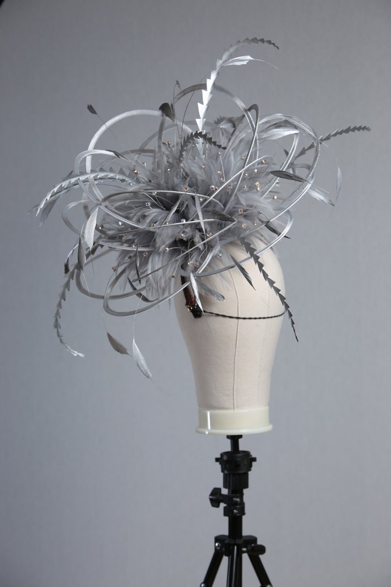 May include: A silver headpiece with feathers and sparkling embellishments. The headpiece is displayed on a mannequin head. The feathers and decorative elements are arranged in a dynamic, upward-sweeping design, creating a dramatic effect. The headpiece is suitable for special occasions.