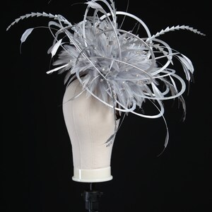 May include: A gray and silver headpiece with feathers and decorative accents. The headpiece is displayed on a mannequin head against a black background. The headpiece features a cluster of gray feathers, silver accents, and small, sparkling embellishments.