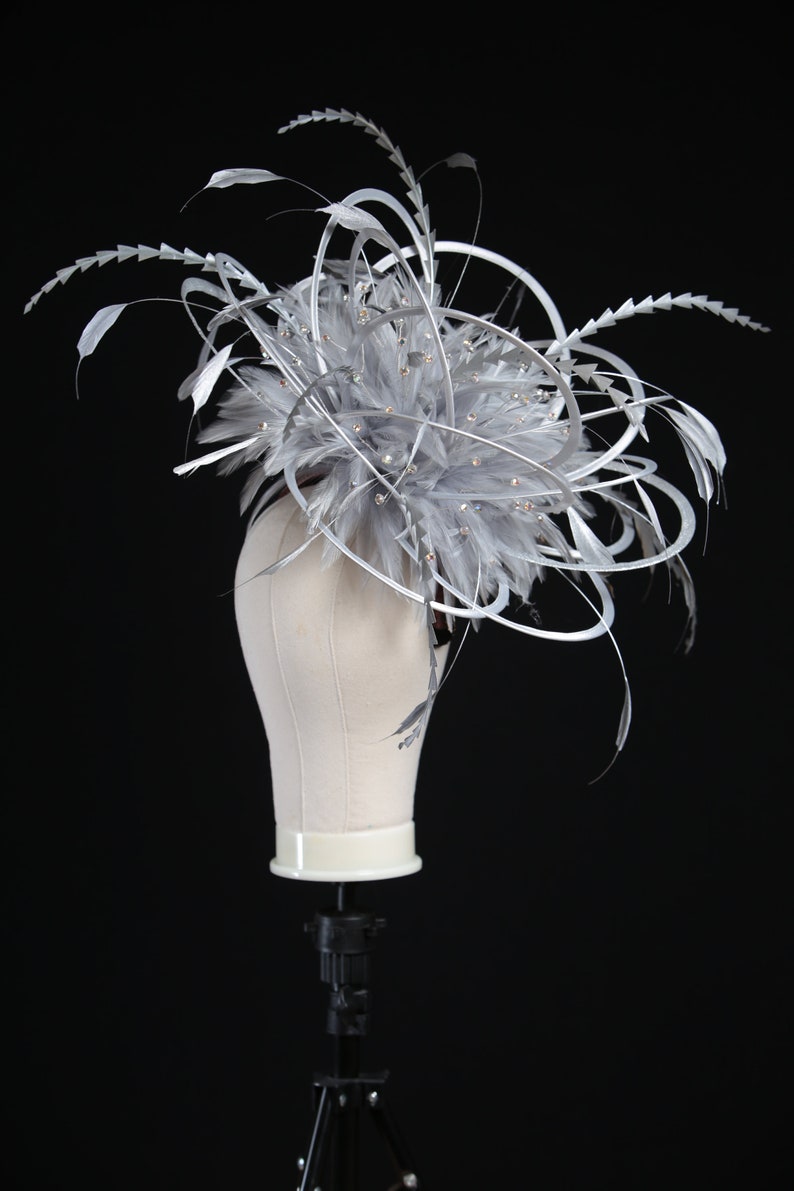 May include: A gray and silver headpiece with feathers and decorative accents. The headpiece is displayed on a mannequin head against a black background. The headpiece features a cluster of gray feathers, silver accents, and small, sparkling embellishments.