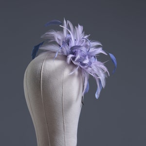 Puede incluir: Un fascinador de plumas lavanda y azul, exhibido en una cabeza de maniquí. El tocado presenta un grupo de plumas lavanda y un acento de cinta, con varias plumas azules que se extienden hacia afuera. El fondo es gris sólido.