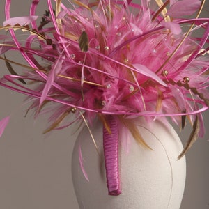 以下が含まれることがあります： 鮮やかなピンク色のヘッドピース。羽、リボン、キラキラ光る装飾が特徴です。デザインは、ピンクの羽の中心的なクラスター、金とピンクのリボン、小さなクリスタルで構成されています。ヘッドピースは布で覆われたフォームに取り付けられています。