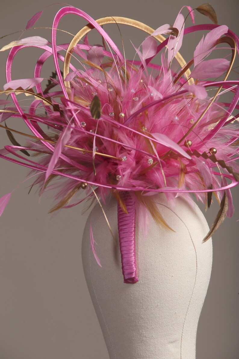 以下が含まれることがあります： 鮮やかなピンク色のヘッドピース。羽、リボン、キラキラ光る装飾が特徴です。デザインは、ピンクの羽の中心的なクラスター、金とピンクのリボン、小さなクリスタルで構成されています。ヘッドピースは布で覆われたフォームに取り付けられています。