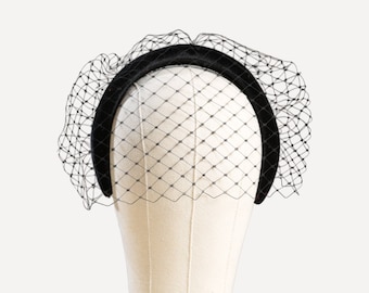 May include: A black headband with a velvet finish, adorned with a delicate black netting veil. The veil is attached to the headband, creating a classic and elegant accessory. The headband is displayed on a mannequin head.