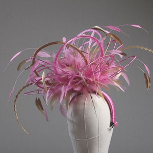 以下が含まれることがあります： ピンクとゴールドの羽根と装飾的なアクセントが付いたヘッドピース。ヘッドピースはマネキンの頭に装着されています。デザインはピンクの羽根、ゴールドのアクセント、ピンクのヘアバンドが特徴です。このアクセサリーは特別な機会に適しています。