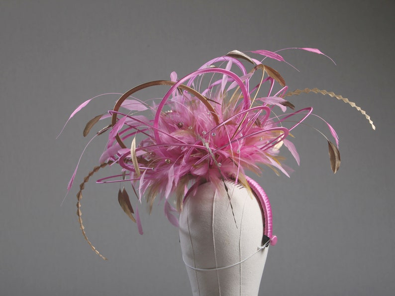 以下が含まれることがあります： ピンクとゴールドの羽根と装飾的なアクセントが付いたヘッドピース。ヘッドピースはマネキンの頭に装着されています。デザインはピンクの羽根、ゴールドのアクセント、ピンクのヘアバンドが特徴です。このアクセサリーは特別な機会に適しています。