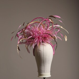 以下が含まれることがあります： ピンクとゴールドのフェミニンな帽子。羽と装飾的なアクセントが特徴です。ヘッドピースはピンクのヘアバンド、ピンクの羽のクラスター、金色の装飾が特徴です。小さな真珠も見えます。