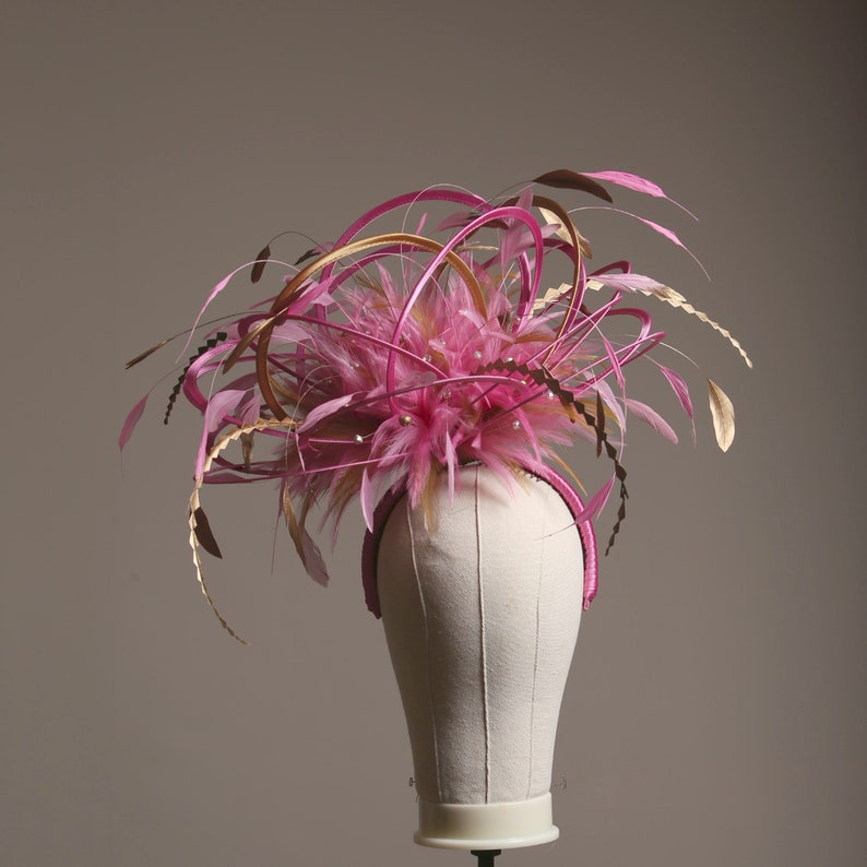 以下が含まれることがあります： ピンクとゴールドのフェミニンな帽子。羽と装飾的なアクセントが特徴です。ヘッドピースはピンクのヘアバンド、ピンクの羽のクラスター、金色の装飾が特徴です。小さな真珠も見えます。