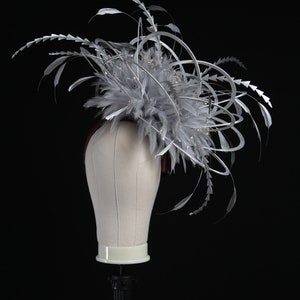 May include: A silver and gray fascinator with feathers and decorative accents. The headpiece is displayed on a mannequin head against a black background. The design features delicate feathers and silver embellishments.