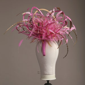以下が含まれることがあります： マネキンの頭に飾られた、鮮やかなピンク色のファシネーター。金色のハローのようなアクセントが特徴です。ヘッドピースは、ピンク色の羽、金色の装飾、小さな真珠のような装飾で飾られています。ヘッドバンドは、お揃いのピンク色です。