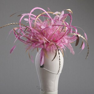 以下が含まれることがあります： ピンクとゴールドのフェザー、リボン、装飾的なアクセントが施されたファシネーター。マネキンの白い頭にグレーの背景で展示されています。デザインは、ピンクの羽と金の装飾の中央クラスターが特徴です。