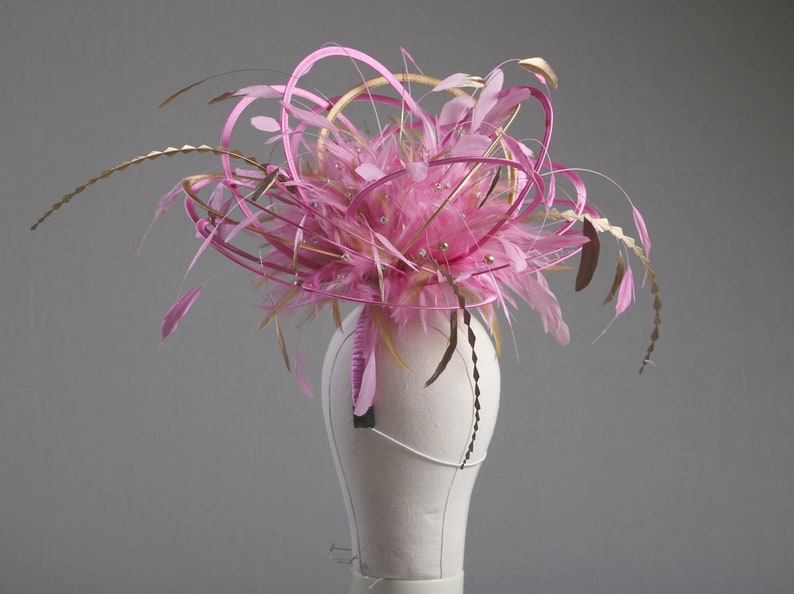 以下が含まれることがあります： ピンクとゴールドのフェザー、リボン、装飾的なアクセントが施されたファシネーター。マネキンの白い頭にグレーの背景で展示されています。デザインは、ピンクの羽と金の装飾の中央クラスターが特徴です。