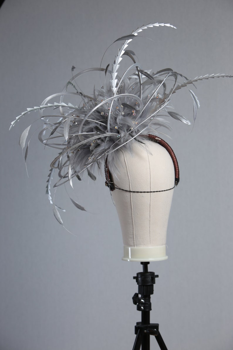 May include: A silver and gray fascinator with feathers and decorative accents. The headpiece is mounted on a mannequin head. The feathers are arranged in a layered design, with a brown headband.