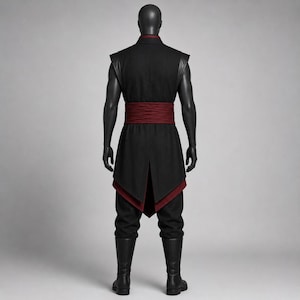 May include: A black sleeveless tunic with a high collar and a wide, textured dark red sash at the waist. The tunic features a layered hemline with a dark red underlayer, paired with black trousers and black boots on a mannequin figure.