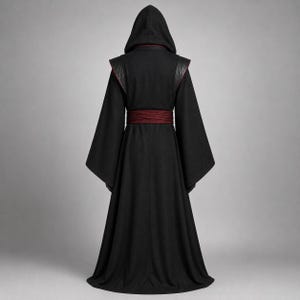 May include: A floor-length black hooded robe featuring wide, flared sleeves and a dark red horizontal waistband. The garment has structured shoulder panels and a pointed hood, presented from a rear-facing perspective against a neutral background.
