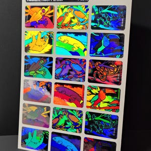 May include: A rectangular sheet of Radiant Rain Forest T-6210 stickers featuring fifteen individual holographic designs. The stickers display colorful illustrations of rainforest animals including frogs, parrots, snakes, sloths, iguanas, and toucans.