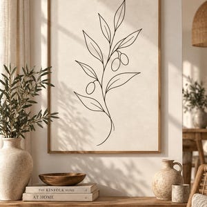 May include: A framed wall art print featuring a black line drawing of an olive branch with leaves and two olives on a light beige background. The artwork is displayed in a thin wooden frame, showcasing a simple botanical illustration.