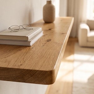Puede incluir: Un estante flotante de madera de color marrón claro con un patrón de grano visible. Sobre el estante hay dos libros blancos apilados con gafas de montura metálica fina encima, y un jarrón cilíndrico alto de cerámica clara colocado más atrás.
