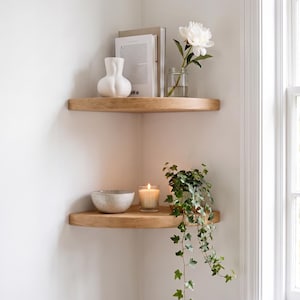 Könnte beinhalten: Zwei Eckregale aus hellem Holz an einer weißen Wand. Das obere Regal enthält eine weiße Keramikvase, ein Buch und eine Glasvase mit einer weißen Pfingstrose. Das untere Regal enthält eine gesprenkelte Keramikschale, eine brennende Kerze und eine hängende grüne Efeupflanze.