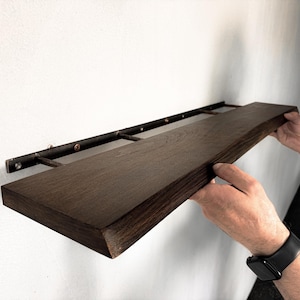 Könnte beinhalten: Ein rechteckiges, dunkelbraunes Holzregal mit sichtbarer Holzmaserung. Das Regal ist an einem schwarzen Wandhalterungssystem aus Metall befestigt, das horizontale Stangen und kleine kreisförmige Befestigungen an der hinteren Kante aufweist.