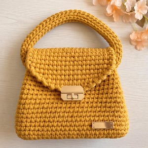 Puede incluir: Un bolso de ganchillo color amarillo mostaza con un asa superior gruesa y redondeada y una solapa. El bolso se asegura con un cierre de presión rectangular de metal dorado e incluye una pequeña placa rectangular de metal dorado en la base.