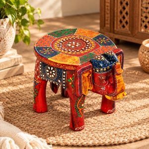 May include: A small, round wooden stool shaped like an elephant, featuring a vibrant, hand-painted design in red, blue, green, and yellow. The surface displays intricate floral patterns, geometric shapes, and small inlaid mirror accents.