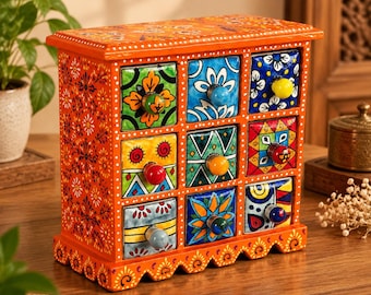 Peut inclure: Un coffre de rangement rectangulaire en bois avec un cadre orange orné de motifs floraux blancs. Il comporte neuf tiroirs individuels, chacun doté d'une façade en céramique peinte à la main et d'un petit bouton rond en céramique unie.
