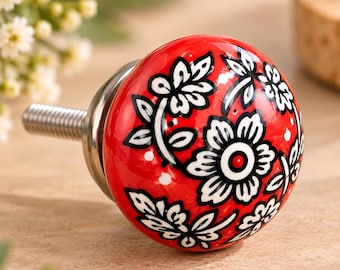 Peut inclure: Un bouton de meuble rond avec une finition rouge brillante orné d'un motif floral noir et blanc. Le bouton possède une base en métal argenté et une tige filetée dépassant à l'arrière pour une installation sur des portes de meubles.