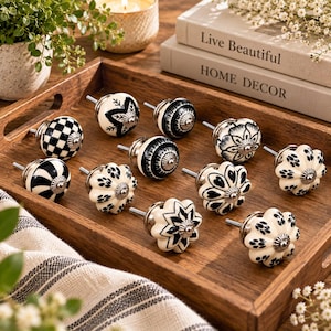 Può includere: Un set di dodici pomelli rotondi in ceramica per mobili con vari motivi in bianco e nero, inclusi disegni a scacchi, floreali, a righe e a stella. Ogni pomello presenta una base in metallo color argento e una vite centrale per il fissaggio.
