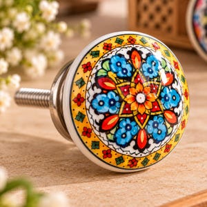 Peut inclure: Un bouton de meuble rond en c&eacute;ramique orn&eacute; d'un motif floral mandala multicolore aux tons jaune, bleu, rouge et vert. Ce bouton pr&eacute;sente une finition brillante et comprend une base &agrave; vis en m&eacute;tal argent&eacute; pour la fixation sur un meuble.
