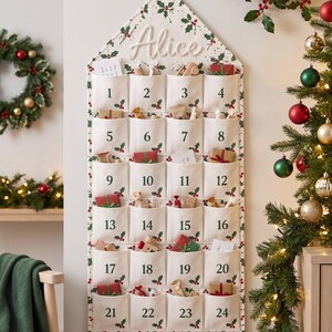 May include: A house-shaped fabric advent calendar with a white background featuring a green holly and red berry pattern. It has twenty-four numbered pockets, each printed with a number from 1 to 24. The name Alice is embroidered in white script at the top.