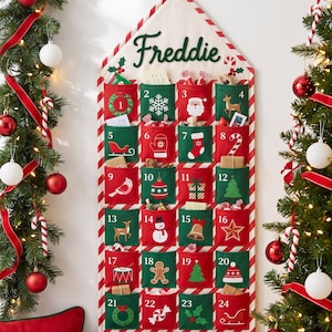 Può includere: Un calendario dell'avvento in tessuto a forma di casa, con il nome Freddie in corsivo verde. Presenta ventiquattro tasche numerate che si alternano tra rosso e verde, decorate con icone natalizie bianche come un pupazzo di neve e un albero.